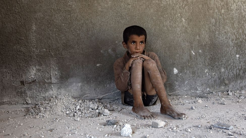 A Palestinian child sits on the bare ground with bare dusty feet and legs, Khan Younis, Gaza, August 2024