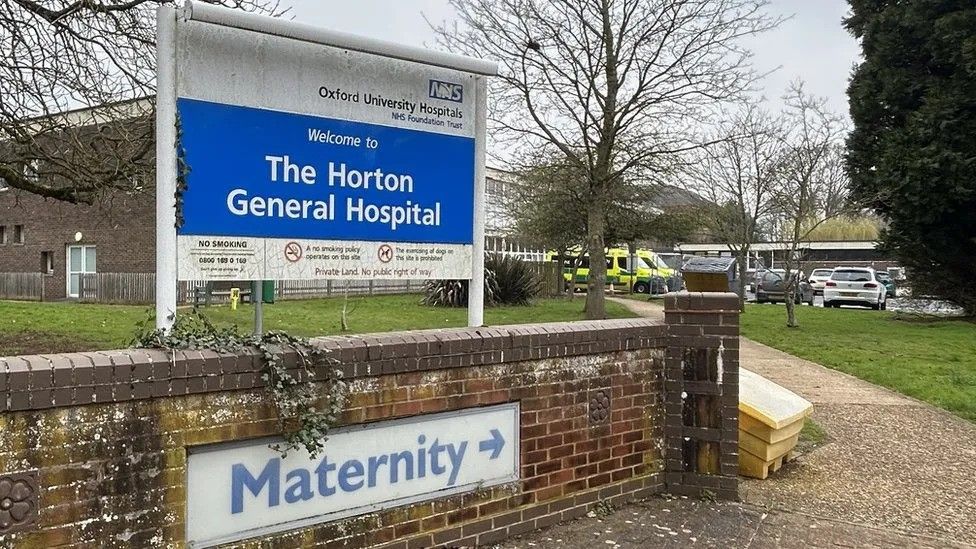 The entrance to the Horton General Hospital. Its sign stands over a low brick wall. A sign for the maternity service is set into the wall. Hospital buildings and a car park are in the distance.