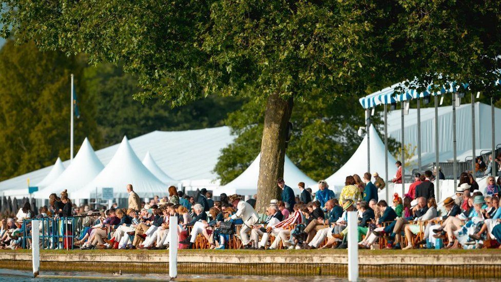 Spectators sat and standing along the River Thames with white marquee roofs behind