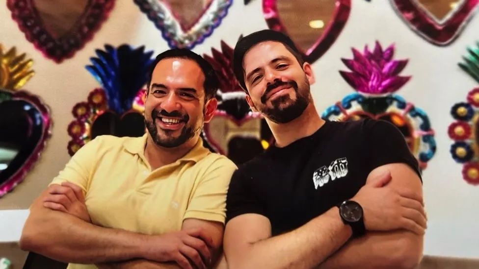 Two men smile at the camera with their arms folded. They are stood in front of a number of mirrors with brightly coloured frames