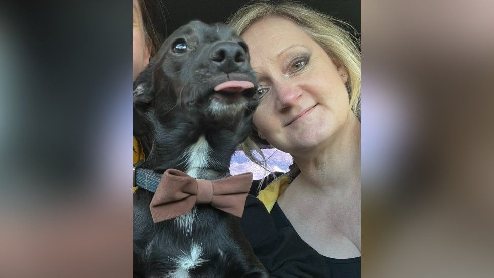 Melanie Gassman, who has blonde hair, next to her dog Harry, who has black wiry fur and is wearing a dark red bow tie