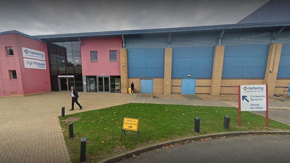 Pink entrance to theatre building with blue and brown exterior walls. There is a small grass patch in the foreground.  A man in a dark suit is walking past.