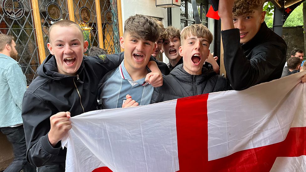 Six England fans celebrate outside a pub holding an England flag after the win over the Netherlands