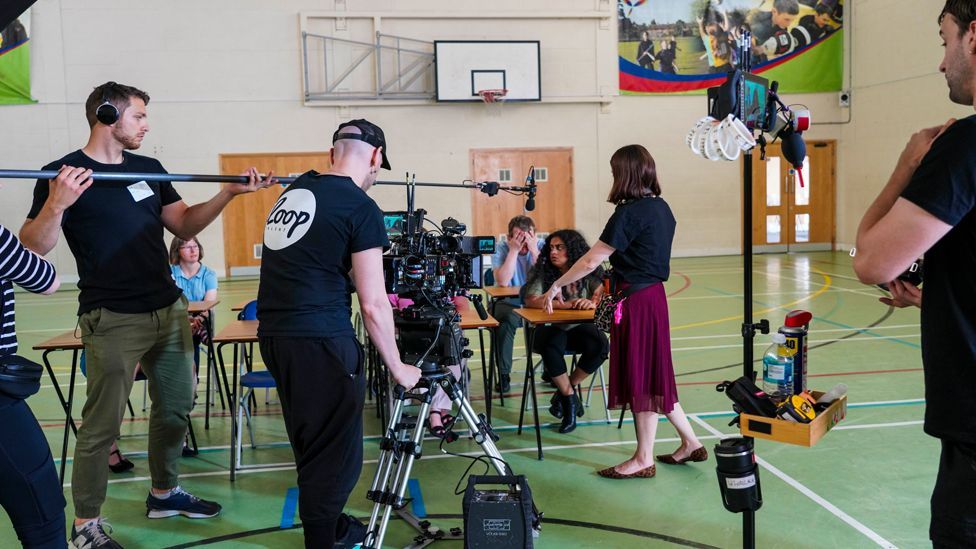 A film set in a gym hall with young people sat at school desks and camera crew filming them