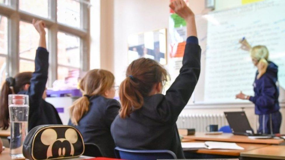 Three school pupils sit with two raising their hands as a teacher writes on a whiteboard in front of them