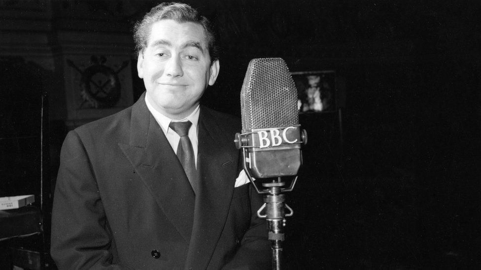 A black and white image of comedian Tony Hancock in a suit, smiling, near a microphone which says BBC on it