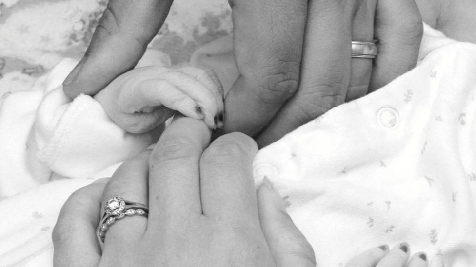 A black and white picture of Wynter being held by her parents. The close-up shows their left hands holding each other and her parents' wedding rings are visible