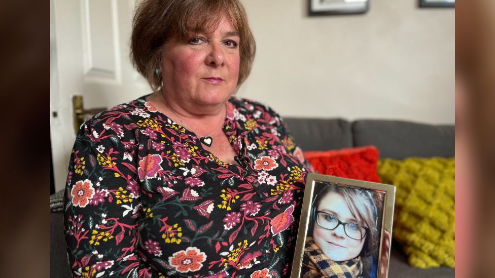 Gina, wearing a black top with floral patterns sitting on her couch, holding up a framed photograph of Cariss.