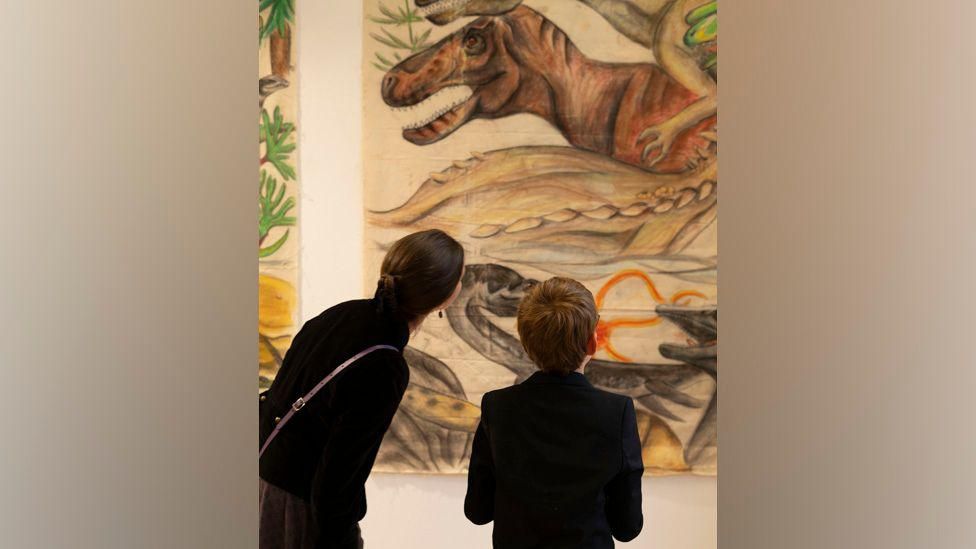 A woman and boy looking at a canvas painting of a T-Rex