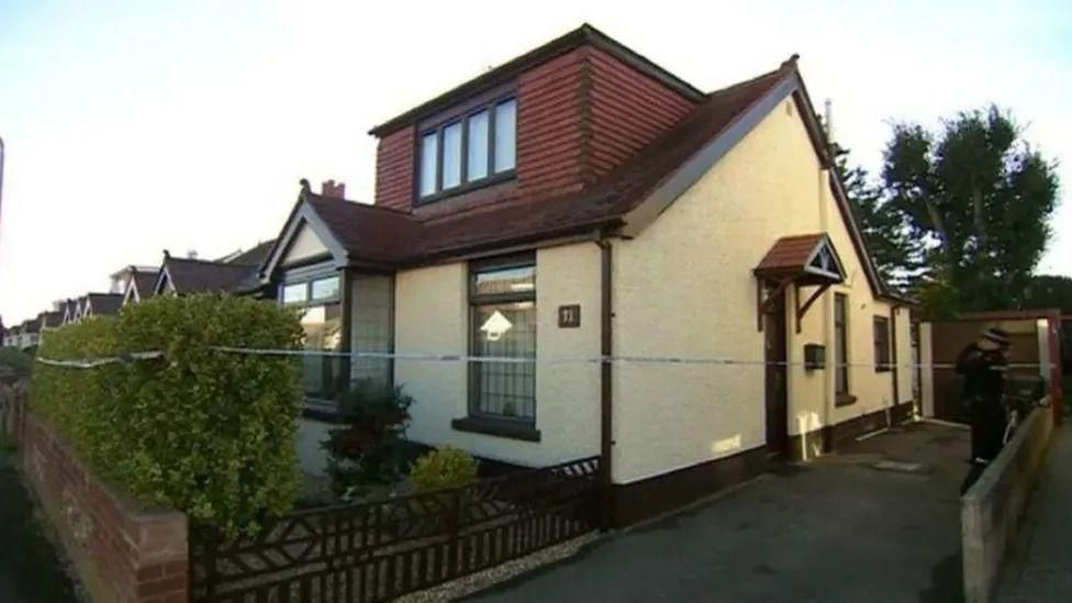 A two-storey house painted cream on the lower half with wooden panelling on the top half. The house is roped off by blue and white police tape.