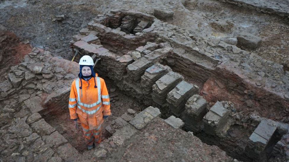 An archaeologist in an orange jump suit with white stripes and a white hard hat standing in the foundations of a Roman tile making unit