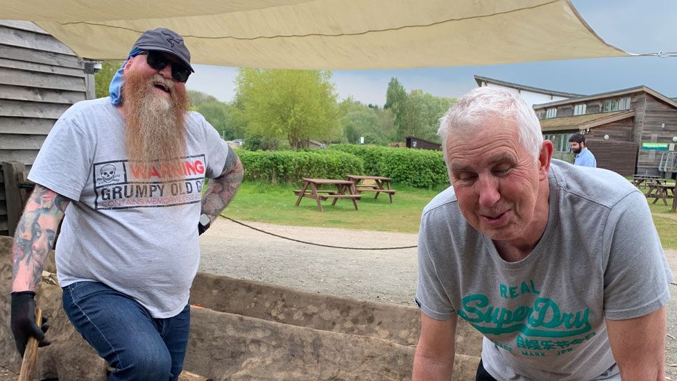 A man with a long beard and wearing a baseball cap laughing alongside another man wearing a T-shirt with his eyes closed and smiling at Stanwick Lakes