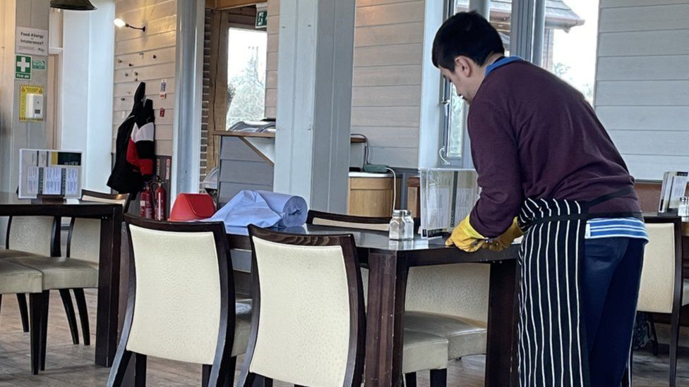 Tommy, a young man with black hair, is wearing a maroon coloured jumper and blue sweatpants with a striped apron and marigold gloves as he leans over a restaurant table to clear it