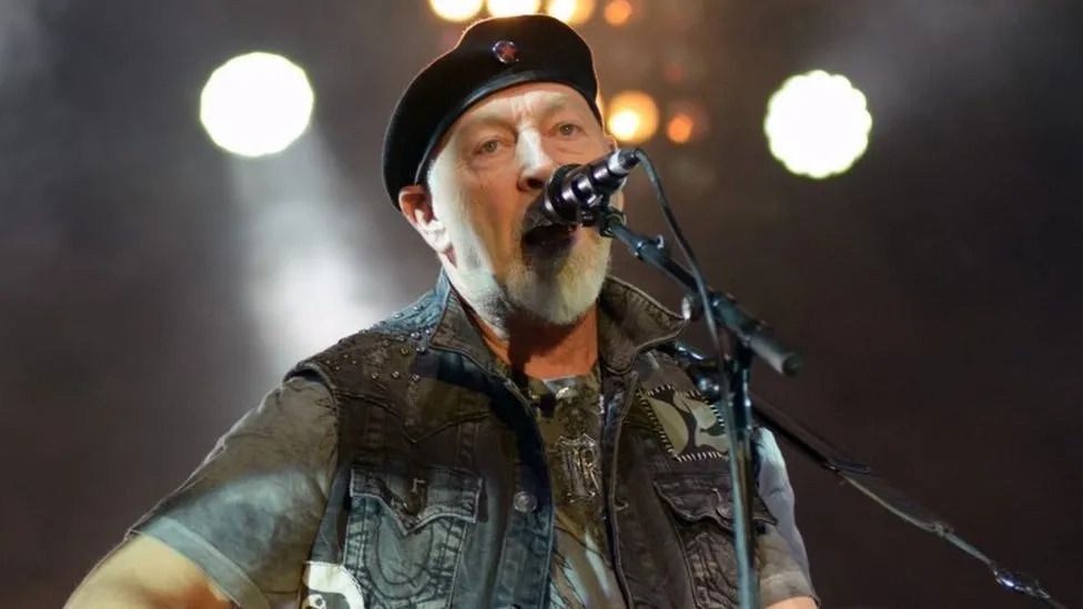 Richard Thompson on stage with a mic and guitar - he is wearing a denim jacket and a beret.