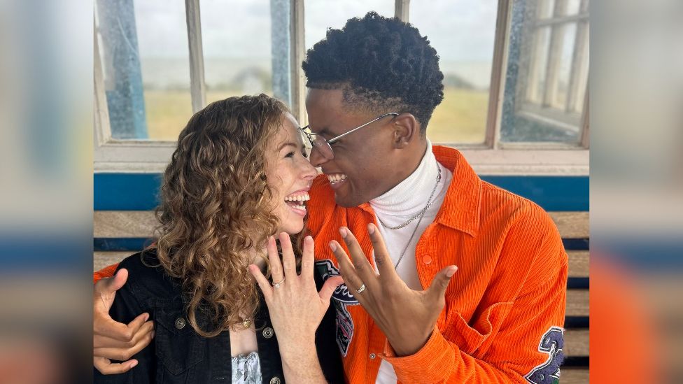 Amy Trigg and Jordan Benjamin, who are sat on a bench while smiling at each other and holding their hands up - both of which have engagement rings on. Amy is wearing a black jacket with a white top and Jordan is wearing an orange jacket with a white roll-neck top.