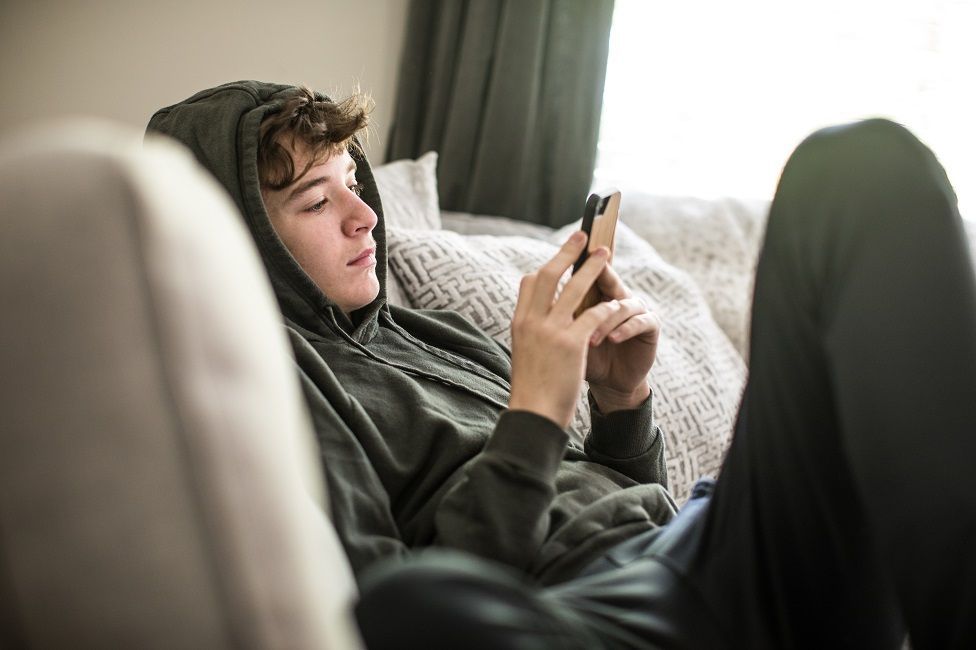 Teenage boy wearing a hoodie sitting on the sofa using smartphone at home