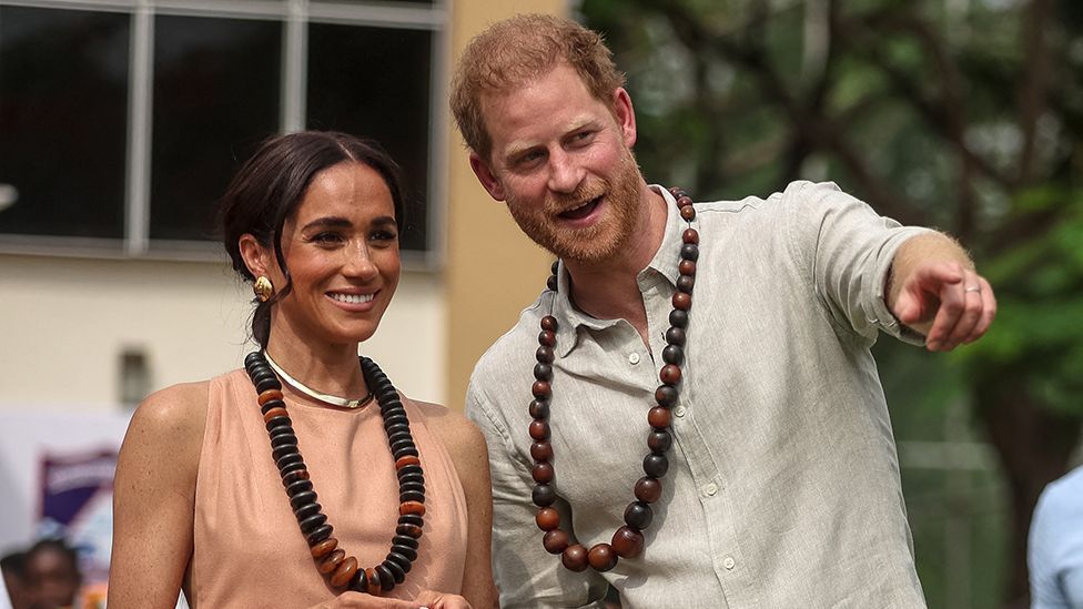 Prince Harry and Meghan, Duchess of Sussex, both smile while wearing large necklaces while Harry points to something out of frame, while in Nigeria in May