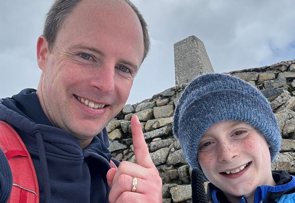 Alex and his Dad Chris at the top of Ben Nevis. Both are smiling at the camera and his dad, Chris, is pointing upwards while wearing a blue jacket with the red armband of a rucksack visible. Alex wears a grey woolly hat and a blue and black coat