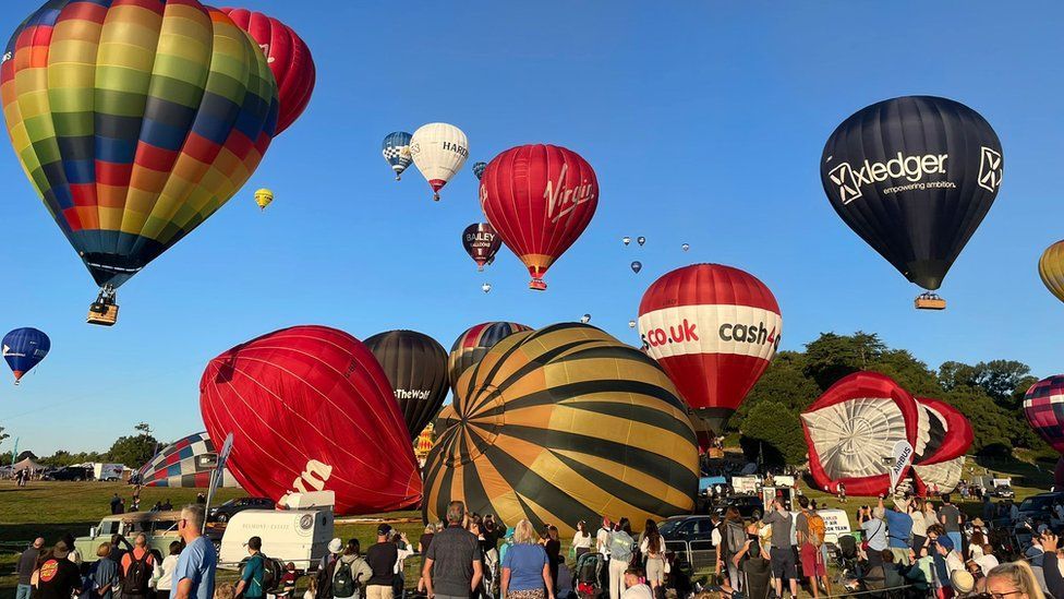 Approximately 22 hot air balloons inflating and taking flight above Ashton Court Estate while hundreds of spectators watch