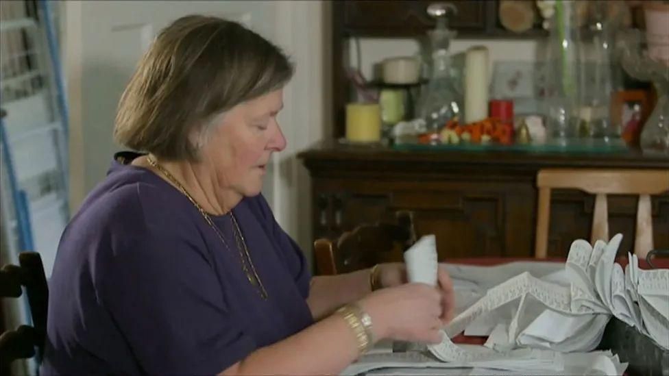 Pam Stubbs sits at a table going through a very long receipt roll. She is wearing a purple top and has a long gold necklace and a gold bracelet. Behind there is a wooden chest of drawers with various glass vases on top.