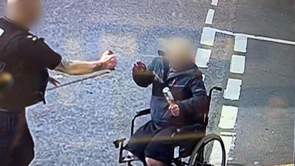 Police officer on left holding a spray towards a man in a wheelchair