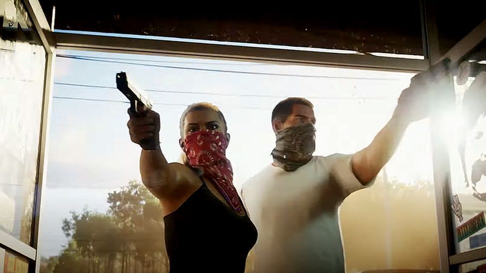 Screenshot shows a male and a female character bursting through the glass front doors of a convenience store. Both have their faces covered by bandanas and each points a pistol into the premises. Behind them the late afternoon sun lights up the background, creating a hazy effect.