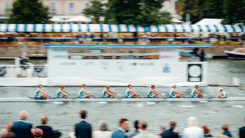 White rowing boat with eight crew members in pale blue racing with blurred crowd in the foreground 