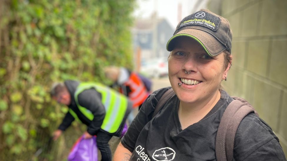 El Clarke, wearing a black T-shirt and a baseball cap, smiling in an alleyway which is being litter-picked.