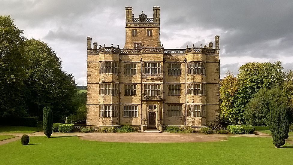 External view of Gawthorpe Hall and gardens - it is a three story, grand building with stairs leading up to it and a large central turret at the back. There are manicured laws and bushes at the front.