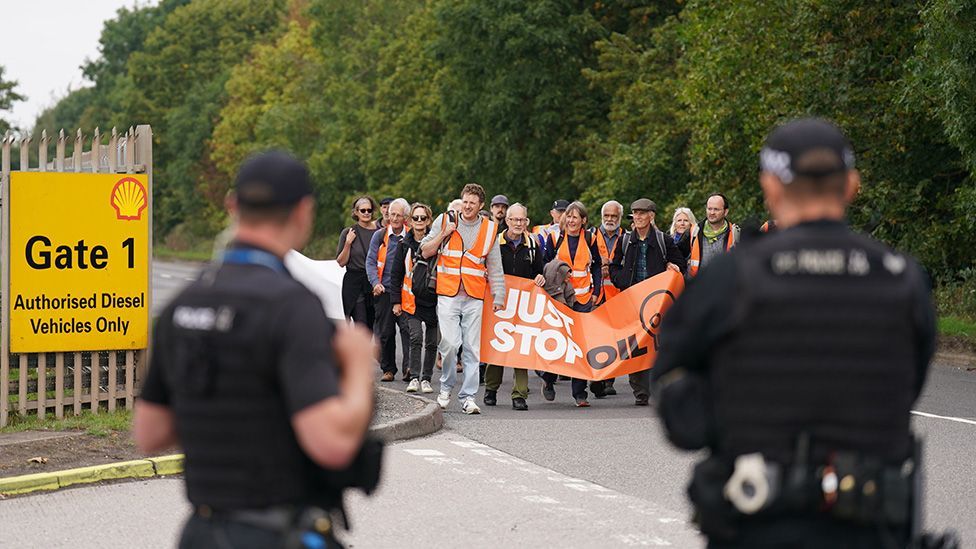 Just Stop Oil protesters approach the gates of Kingsbury oil storage depot, watched by security guards