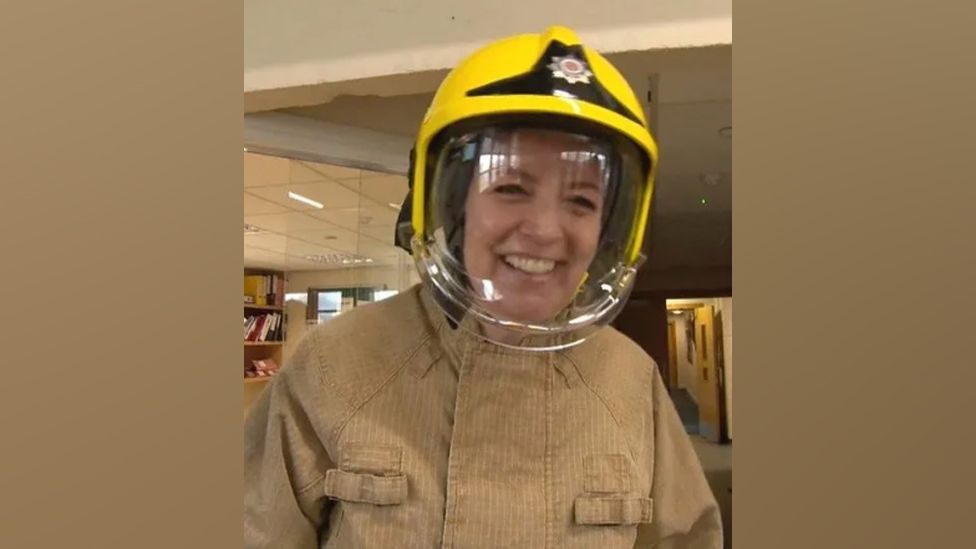 Beccy Bar wears a firefighter's uniform and is smiling at the camera. She is wearing a fire fighters helmet.