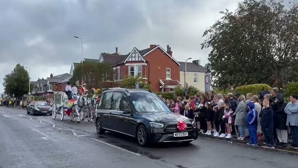 Elsie Dot Stancombe's funeral cortege passes through Birkdale on its way to the church. A hearse drives in front of a white carriage being pulled by white horses, as hundreds line the street. 