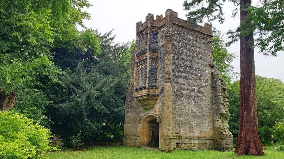 A crenelated stone tower with a large arched doorway. Above the door are two ornate bay windows, one above the other. The tower has broken stones on one side suggesting it was part of a larger building. It is surrounded by trees.