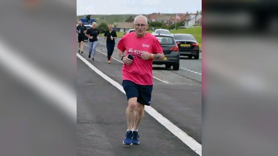 A man with white shaved hair running at the side of a road with cars travelling down the side. He's wearing a pink Blaydon Races T-shirt and holding his phone. People can be seen running behind him.