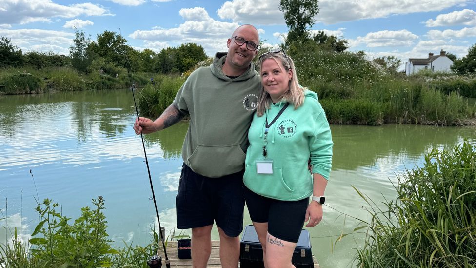 A man and a woman wearing Recovery Rods hooded tops and black shorts stand in front of a fishing lake. The man has a fishing rod in one hand and has his other arm around the woman's back as they smile towards the camera