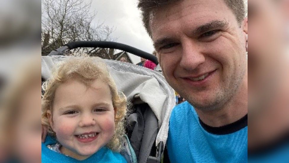 Luke Bakewell smiles next to his daughter Sophie in a pushchair