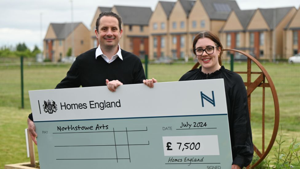 A man and a woman smile at the camera as they hold up a large cheque to Northstowe Arts for £7,500. They are photographed outside with a row of residential houses behind them. 