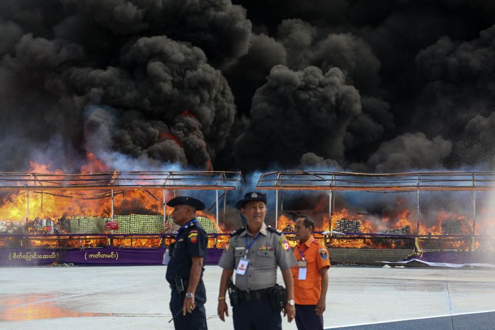 Three officials in uniform stand in front of a burning pile of seized illegal drugs behind a wire fence during a destruction ceremony in Yangon on June 26, 2024.