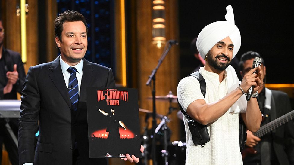 Two men on stage. Host Jimmy Fallon on the left wearing a dark three piece suit with a blue striped tie, and artist Diljit Dosanjh on the left wearing a light coloured outfit and turban, holding a black microphone 