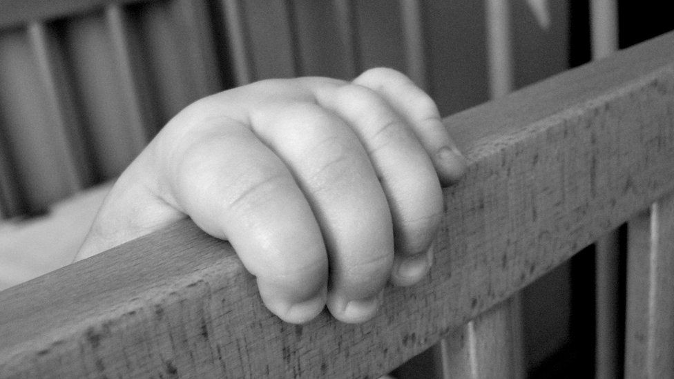 A baby's hand holds on to the side of a cot
