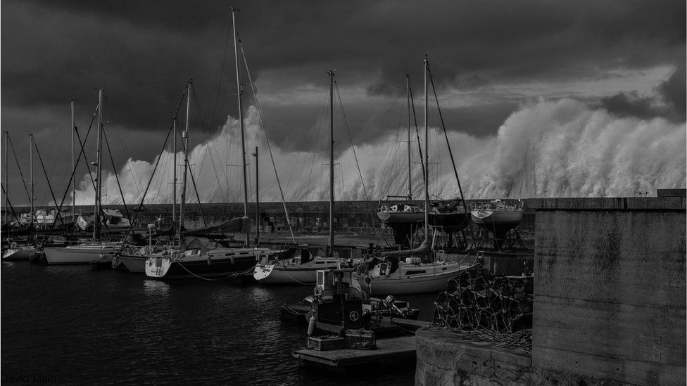 A black and white shot of boats in a harbour. Massive waves crash up and spray over the harbour wall.