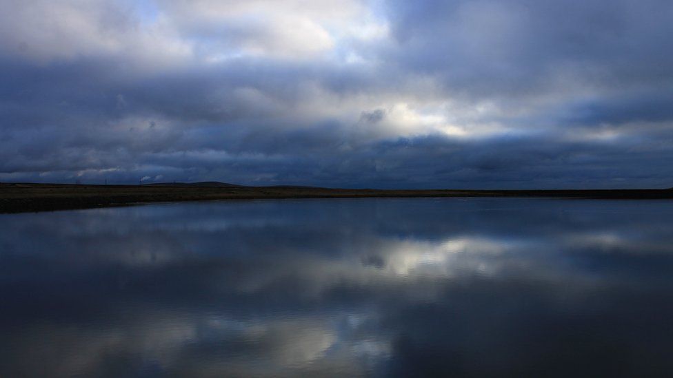 Blue sky and clouds are reflected in a calm blue water below. Land and gentle hills can be seen on the horizon.