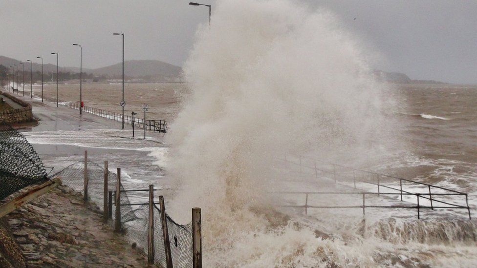 A huge sea spray comes over the sea wall, as high as a tall lamp-post.
