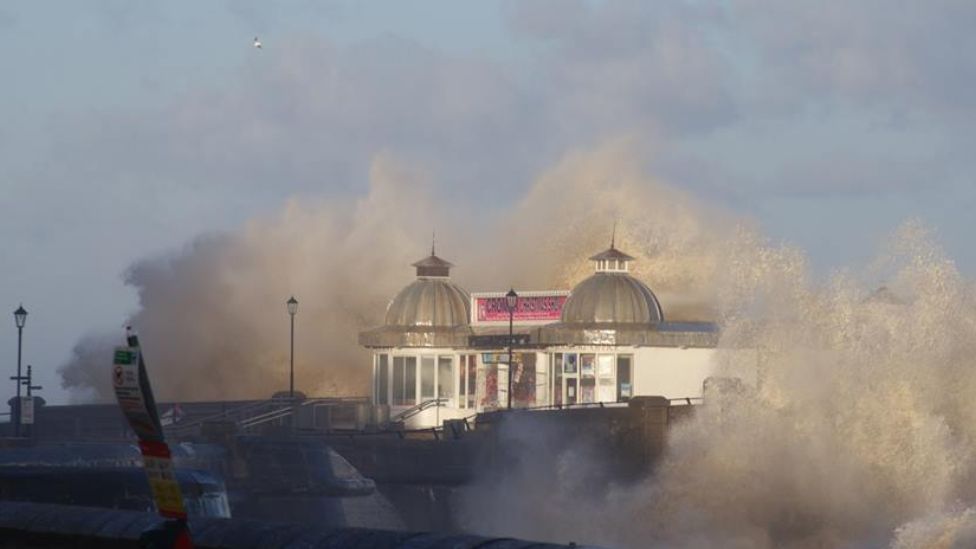 A building on a pier is engulfed by spray from a huge wave.