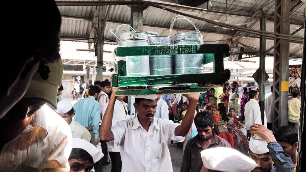 Dabbawala carries a tray of tiffin tins through a crowded railway station.