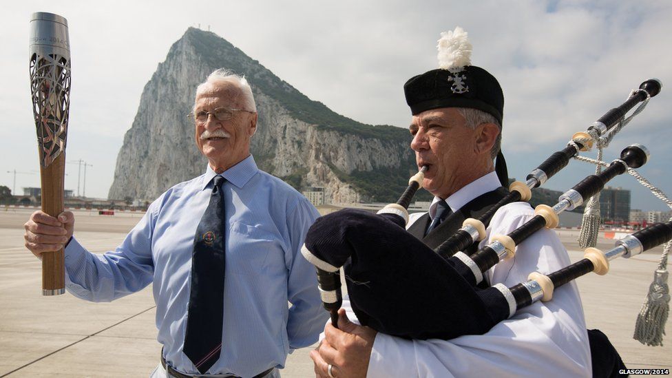 An elderly gentleman holds the Queen's Baton next to a man playing bagpipes. The Rock of Gibraltar is in the background.