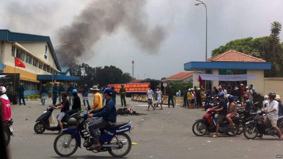 Commuters drive past as riot police stand guard outside a burning factory building in the Vietnamese southern province of Binh Duong on 14 May, 2014