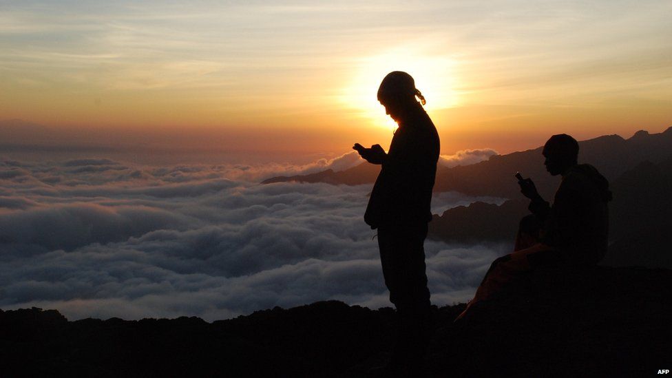 Tanzanian porters and guides stand on a rock halfway up Mount Kilimanjaro at sunset on 21 September 2014 trying to get a mobile phone signal to call their wives