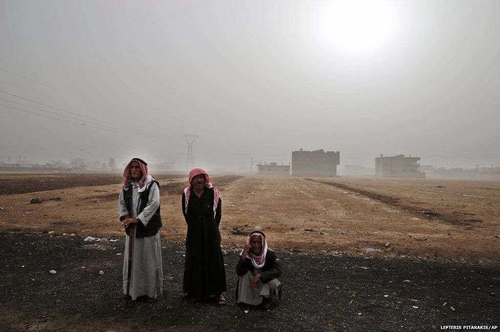 Syrian Kurdish refugees who fled Kobane, stand and the other sits outside the grounds of a refugee camp in Suruc,