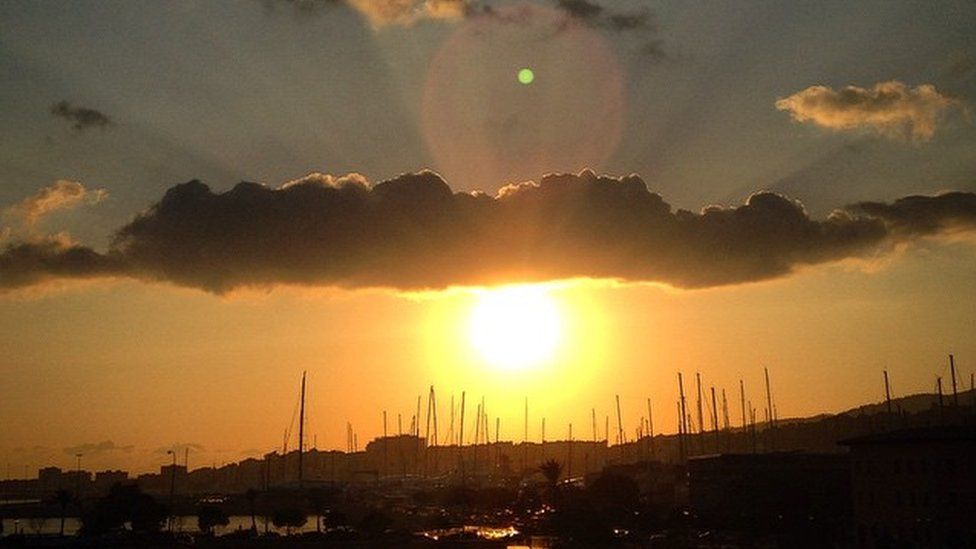 A cloud over the top of the sun as it goes down behind boats in a harbour.